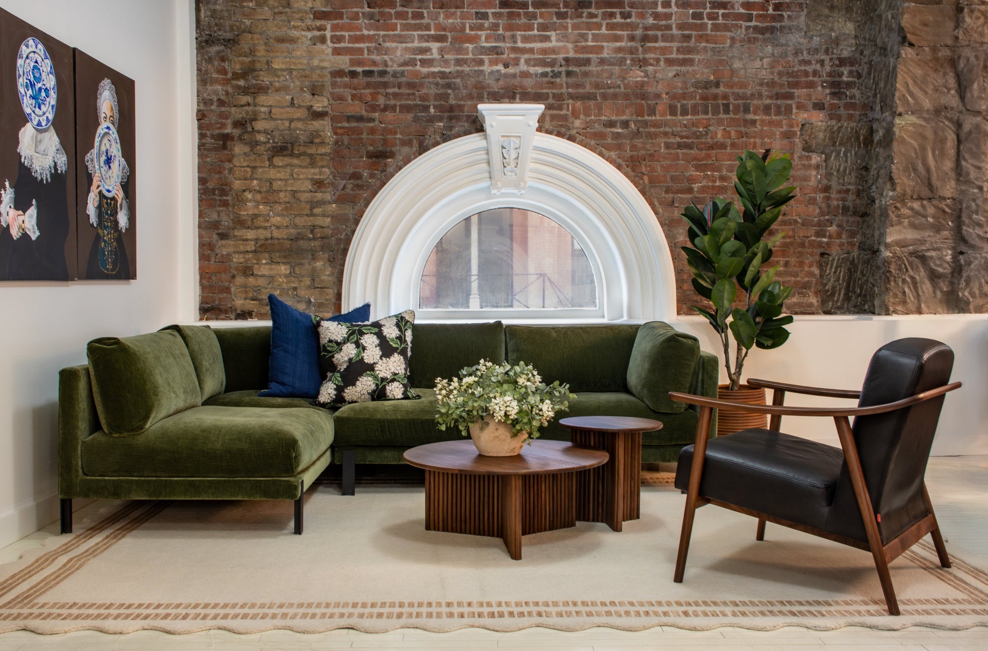 A styled interior scene showing a green velvet sectional sofa, a black leather armchair, round wooden coffee tables, a patterned rug, a brick wall, and a white arched window. No people or animals are present, but this is not a clean isolated product shot because it is set in a real room environment.
