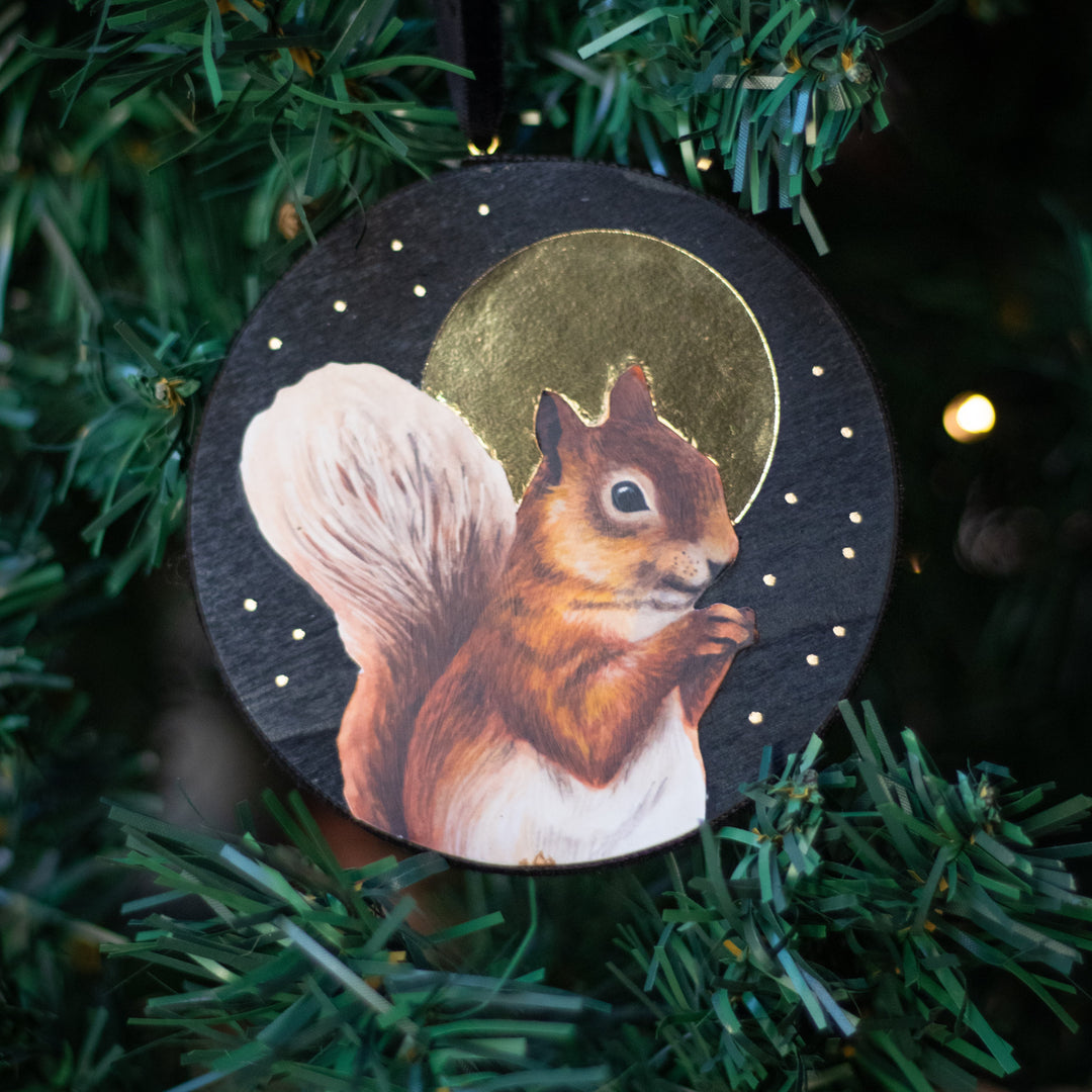 A round wooden Christmas ornament featuring a painted brown and white squirrel in front of a gold moon on a black starry background, hanging on a green artificial Christmas tree.