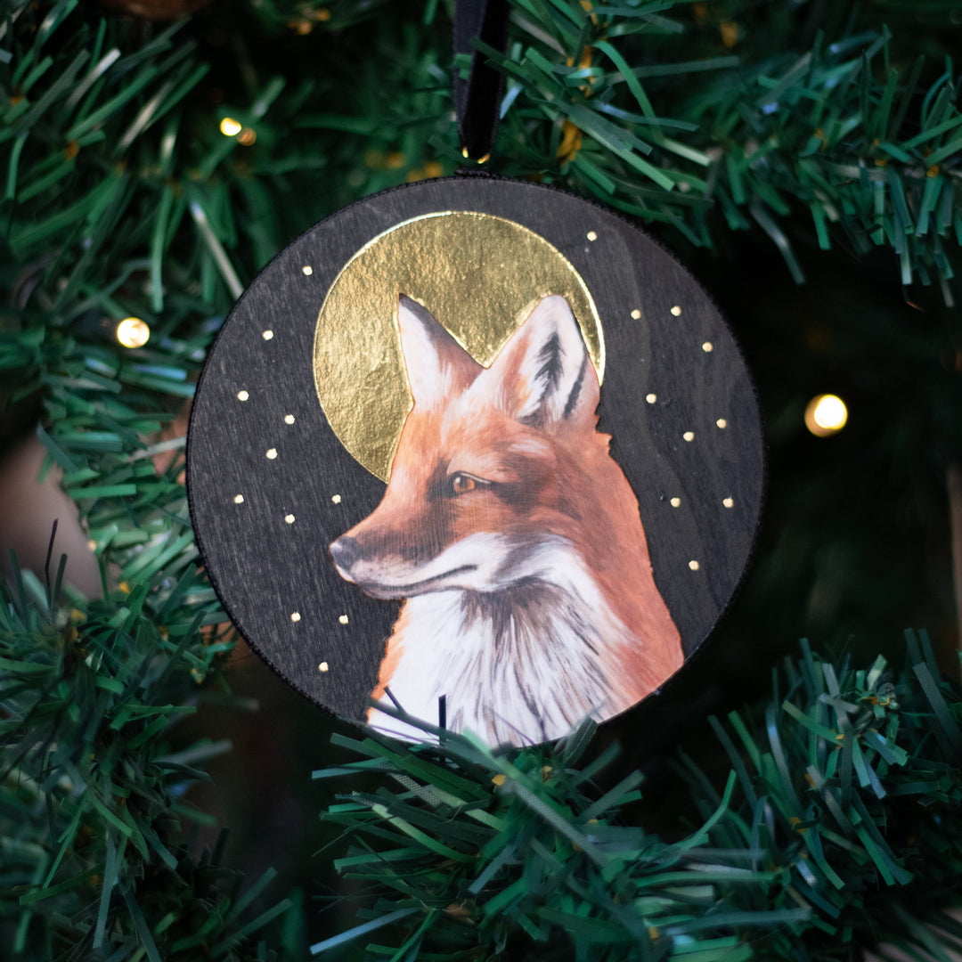A round wooden Christmas ornament featuring a painted fox in profile against a gold crescent moon and starry sky, hanging on a green artificial Christmas tree.