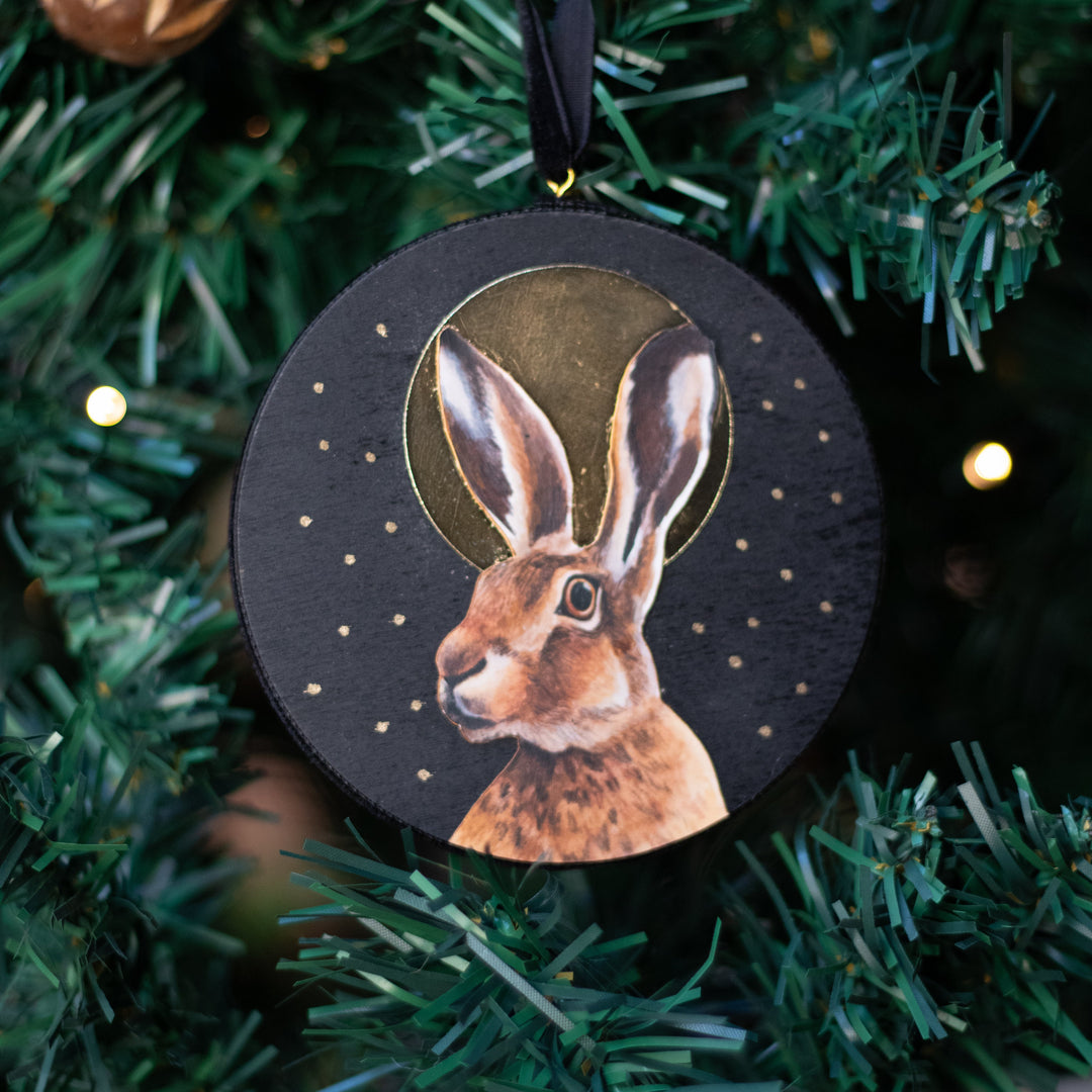 A round black Christmas ornament featuring a painted brown hare with a gold halo, hanging from a black ribbon on a green Christmas tree with warm lights.