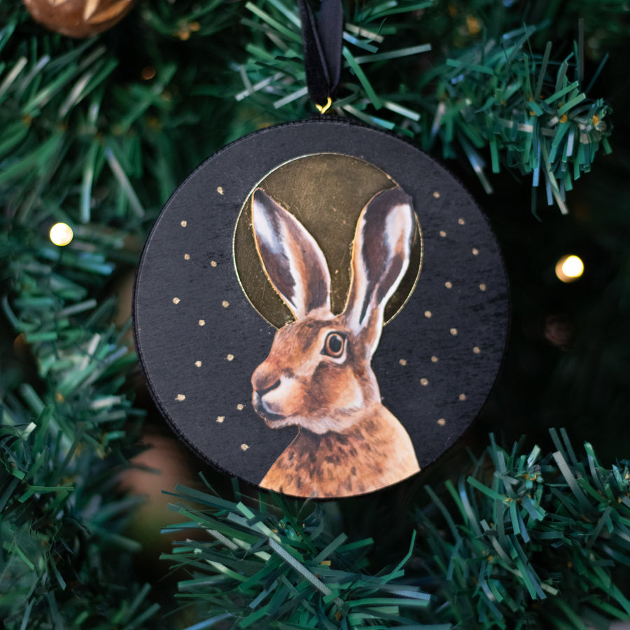 A round black Christmas ornament featuring a painted brown hare with a gold halo, hanging from a black ribbon on a green Christmas tree with warm lights.