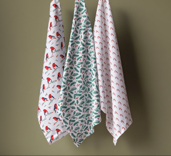Three patterned kitchen towels hanging from a wooden hanger against an olive green wall, with a red box of towels at the bottom right.